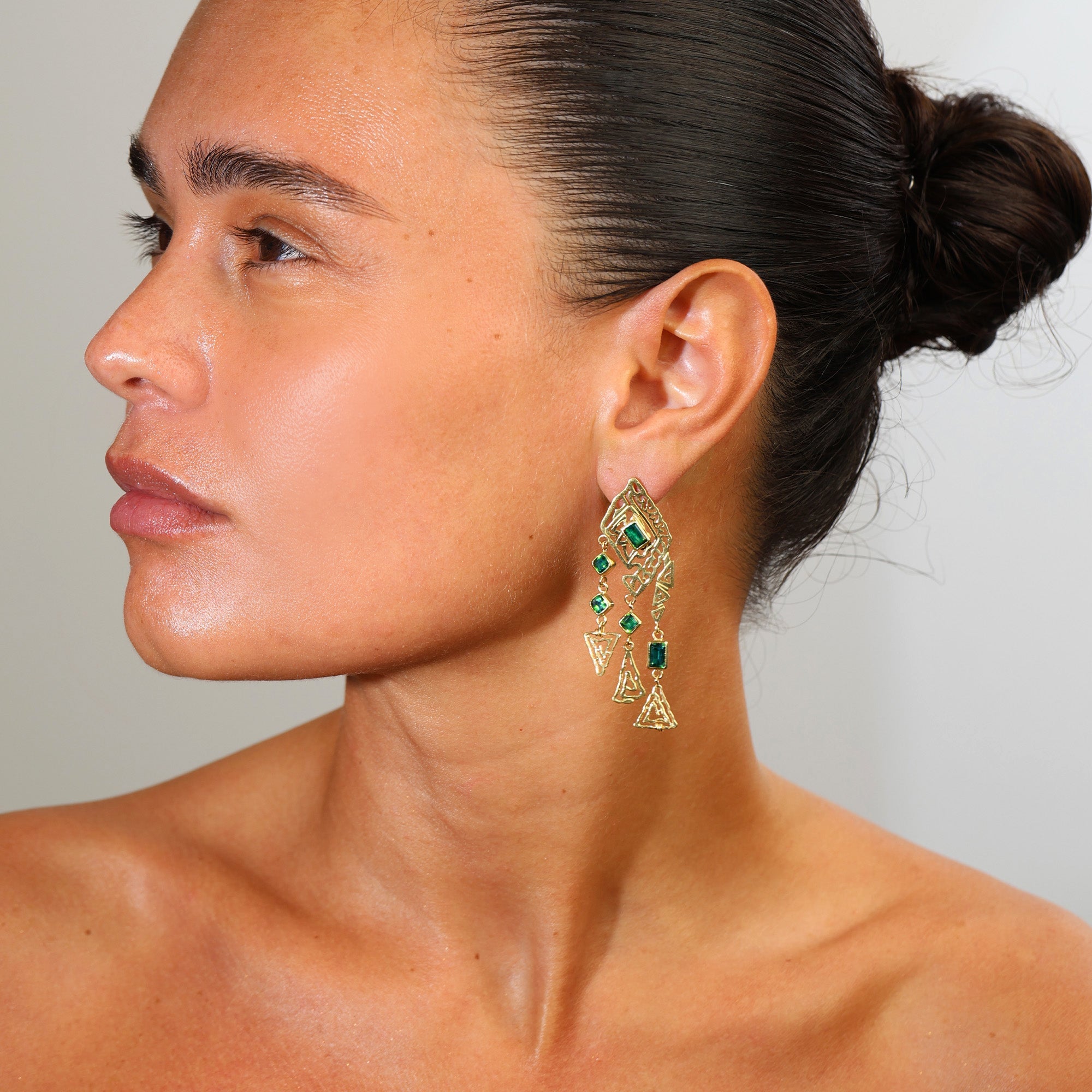 Close-up of a woman wearing gold earrings with green gemstones against a neutral background