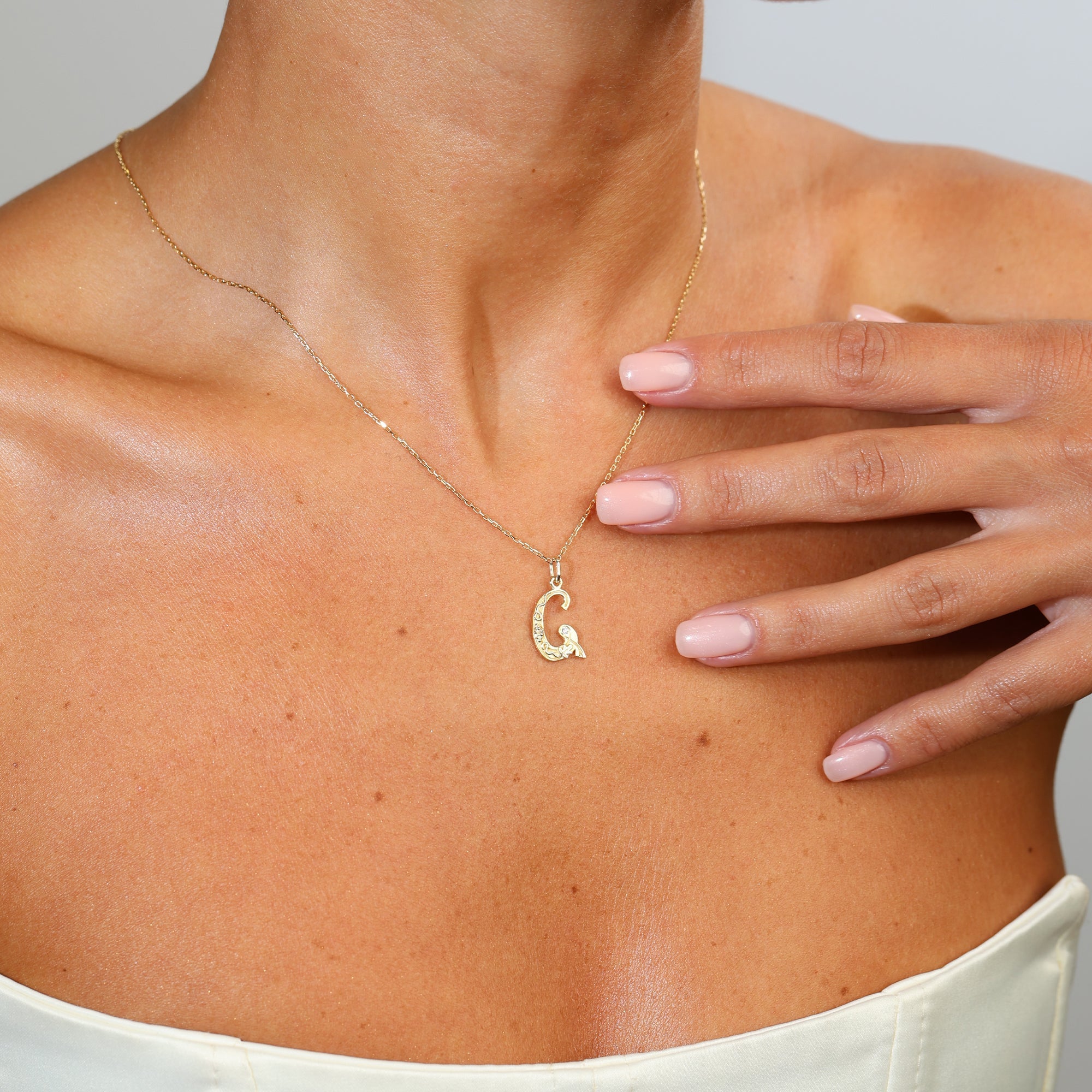 Gold necklace with a 'G' pendant worn by a person, with a hand touching the chest.
