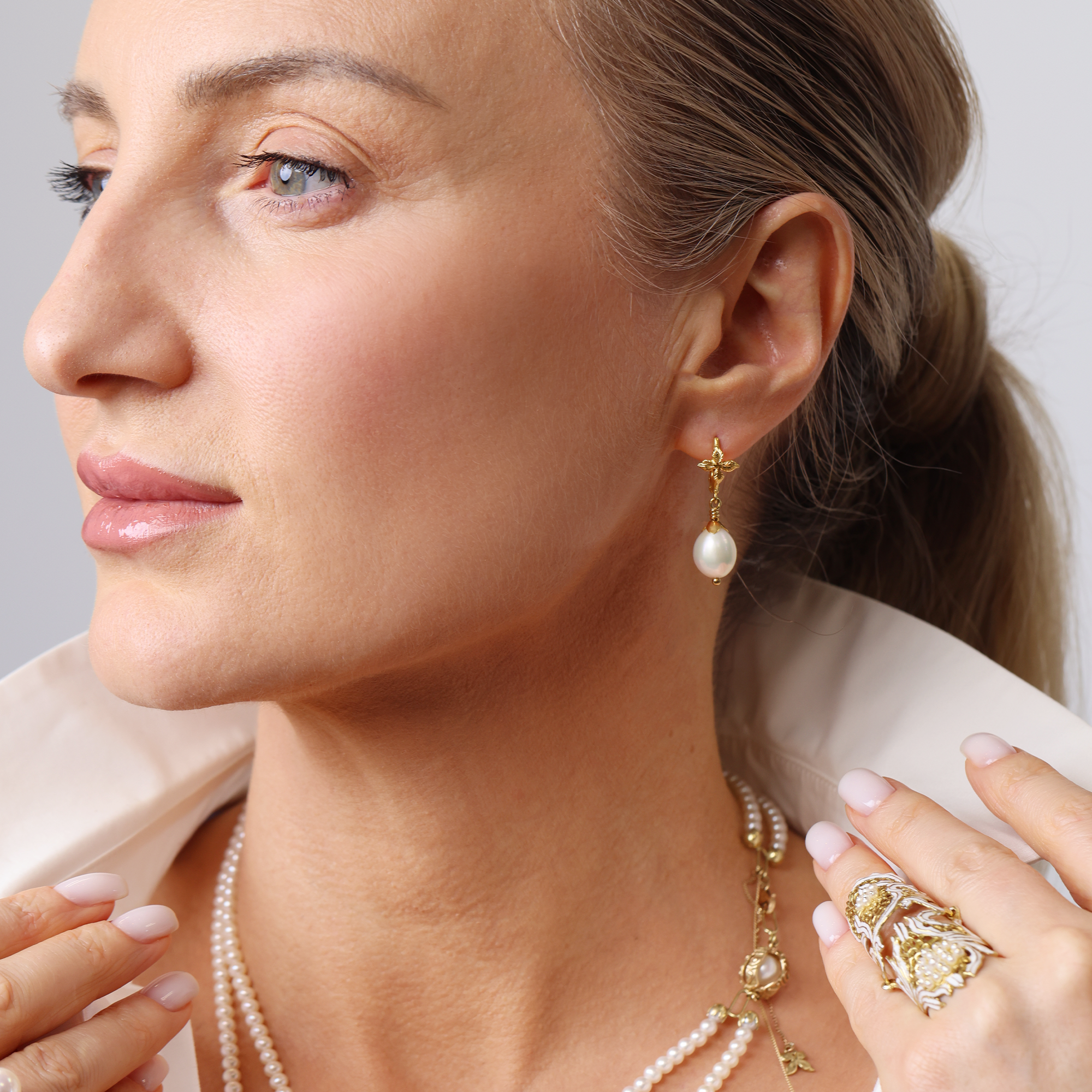 Close-up of a blond woman wearing pearl earrings and a necklace against a neutral background.